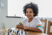 Smiling happy smart African American girl sitting at table with chessboard and learning chess game